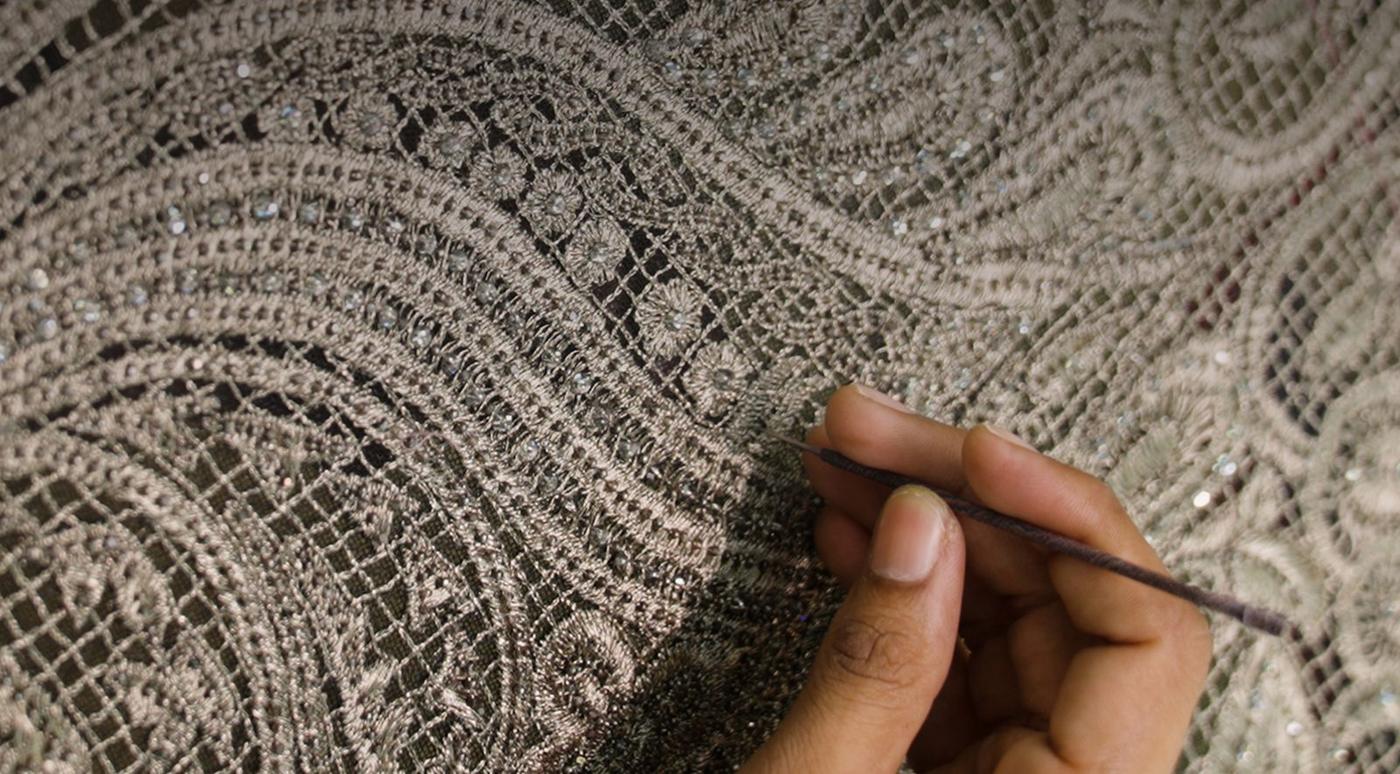 A hand drawing a hooked needle through dense pale thread work set with small stones