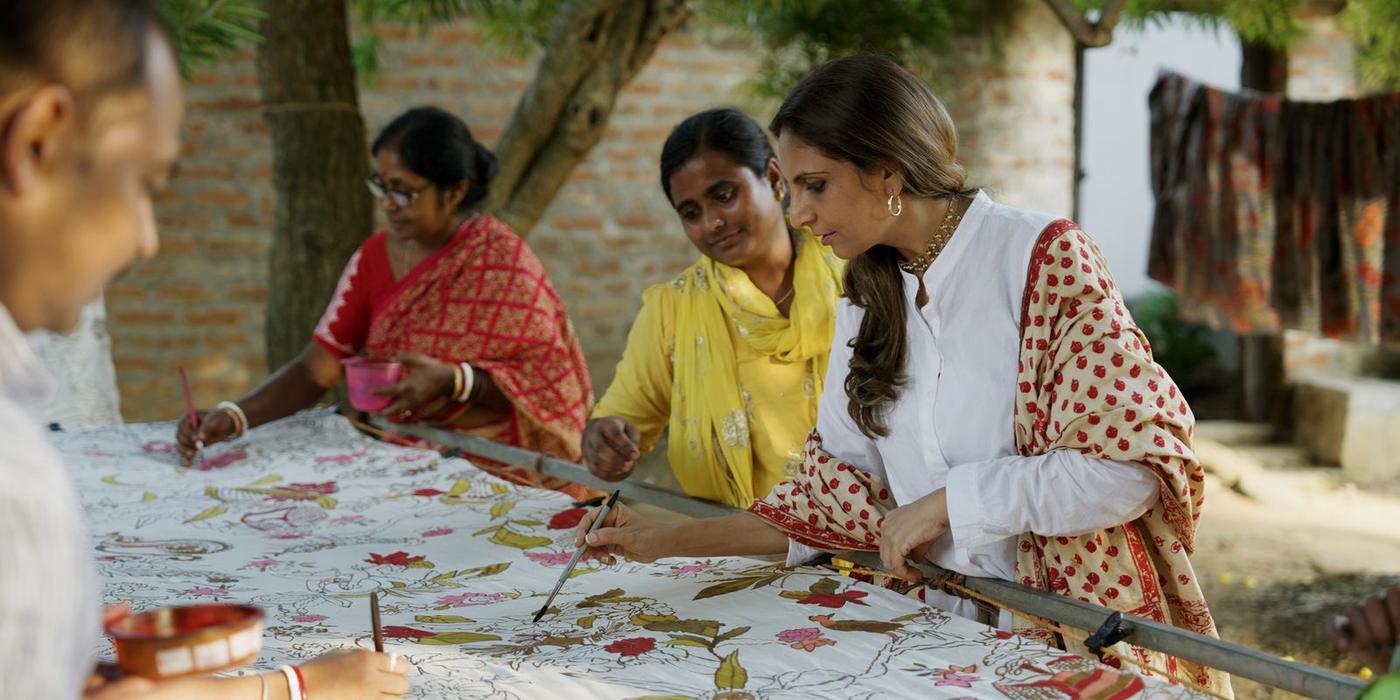 Artisans working side by side over a shared length of cloth, outdoors at the atelier